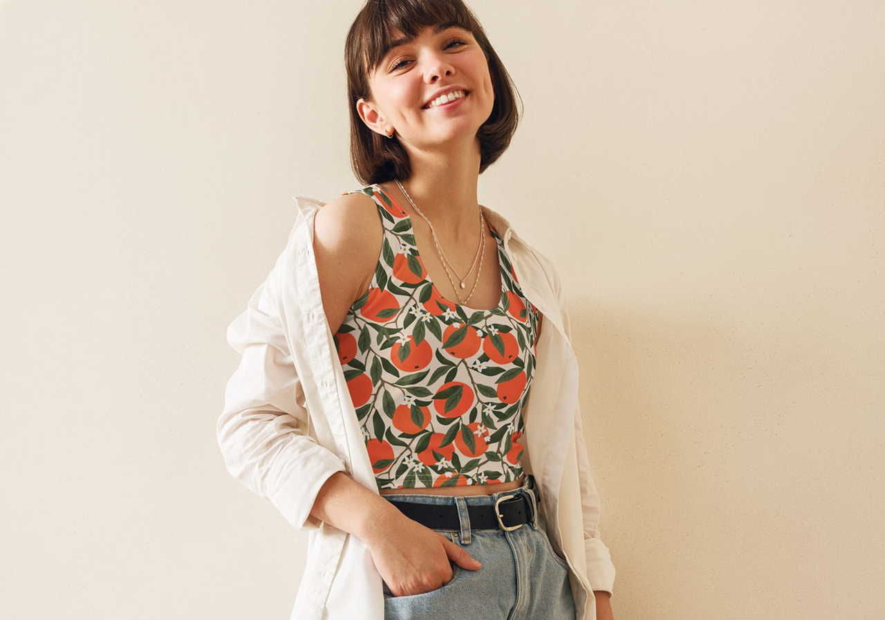 A smiling woman wearing a colorful, floral tank top under an open white shirt, paired with jeans. The background is a sand-yellow.
