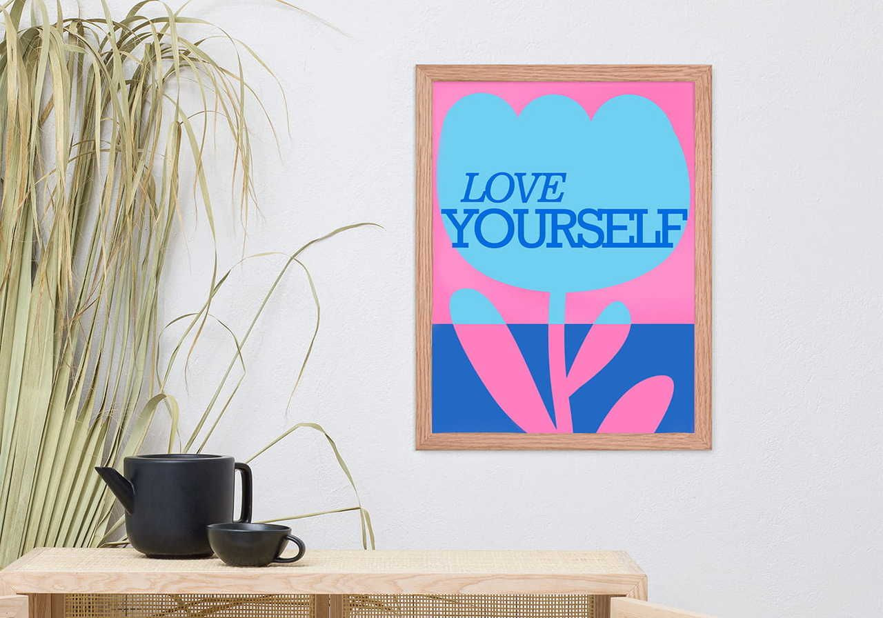 Framed artwork on a white wall with the phrase "Love Yourself" in blue on a blue and pink flower. Black teapot and cup on a light wooden table.