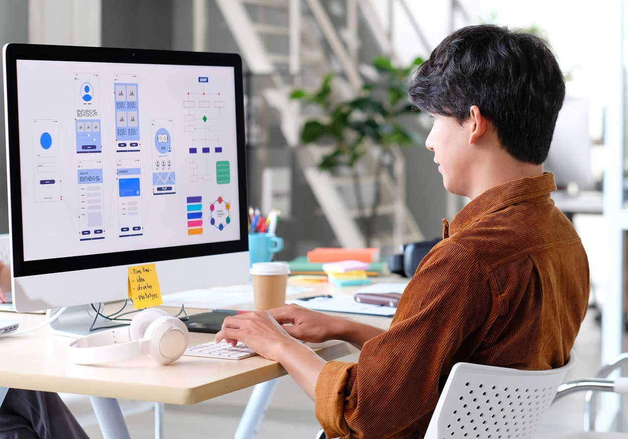 A person in a brown shirt sits at a desk working on a computer displaying website design layouts.