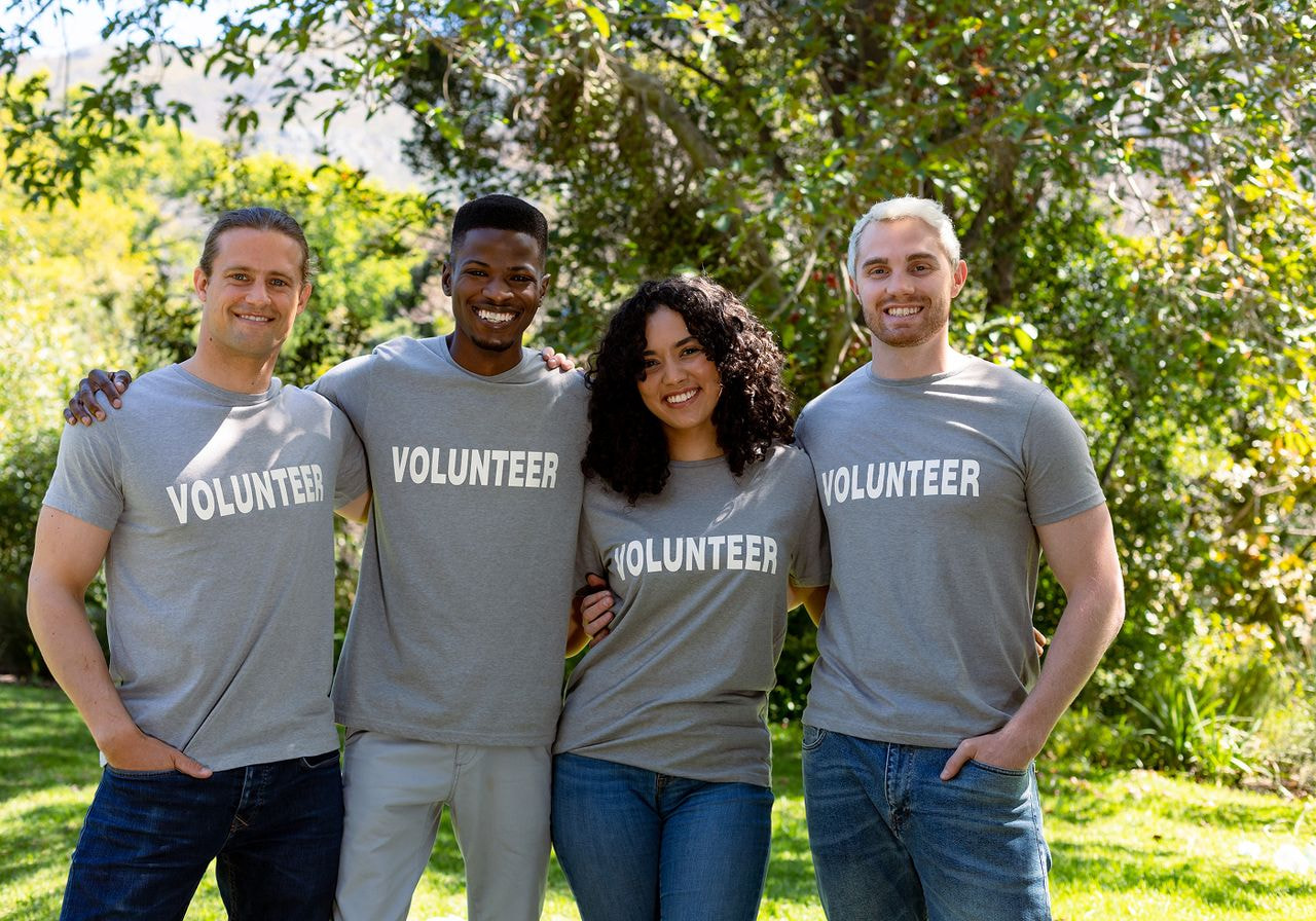 Four smiling volunteers wearing gray 'Volunteer' t-shirts standing outdoors together.