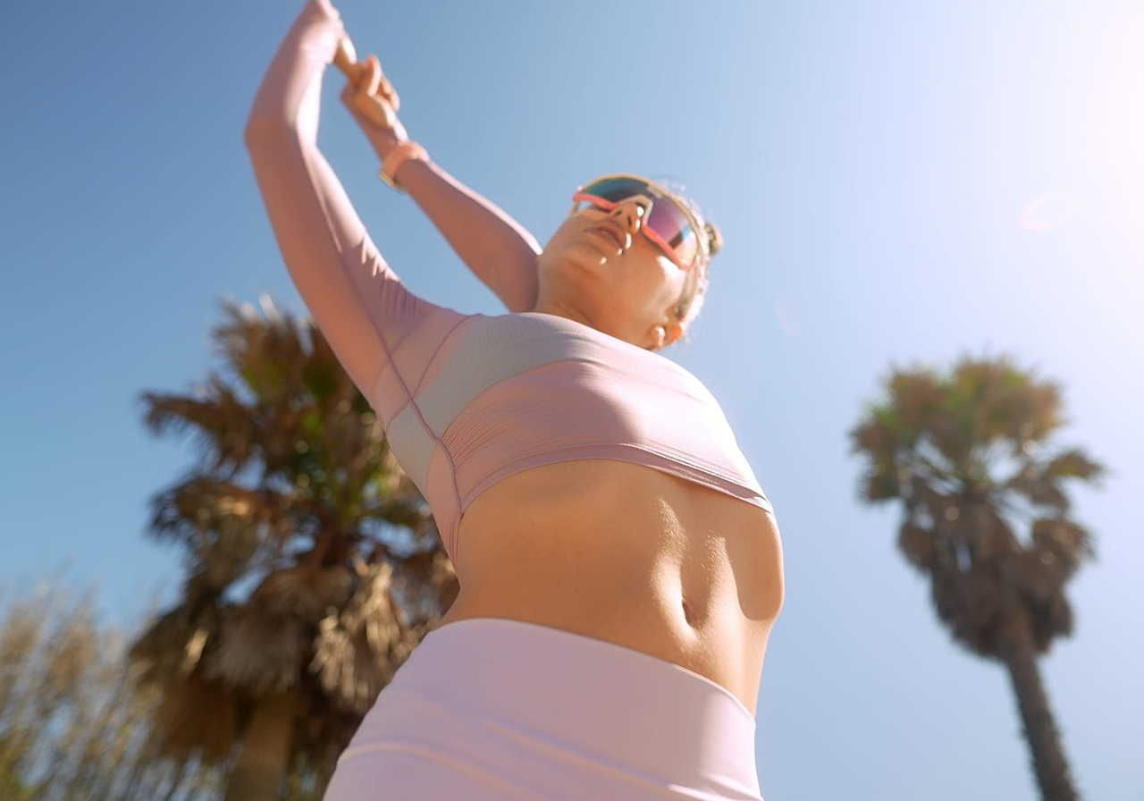 A woman stretching in a light pink outfit under a clear blue sky, with palm trees in the background.