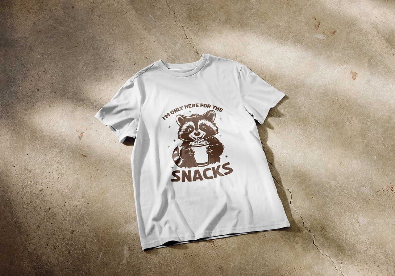 White t-shirt on a concrete floor with a playful cartoon raccoon holding snacks. Text above and below reads, "I'm only here for the snacks".