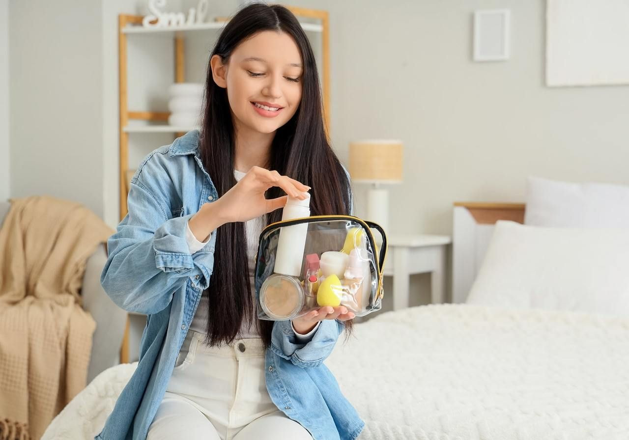 A young woman in a blue denim jacket and white pants smiling while organizing beauty products in a clear travel bag.