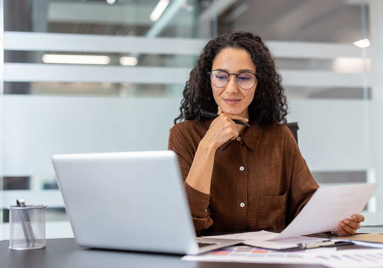 A woman in glasses sits at a desk, holding a document and pen, thoughtfully looking at a laptop. Office setting.
