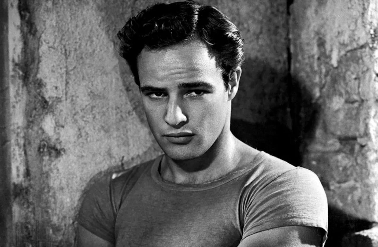 Black and white photo of a man standing against a textured wall, wearing a fitted T-shirt. 