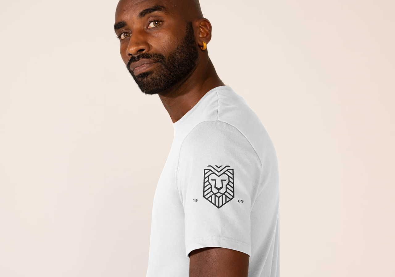 A man in a white t-shirt with a geometrical lion logo on the sleeve, looking over his shoulder. The background is a soft beige.