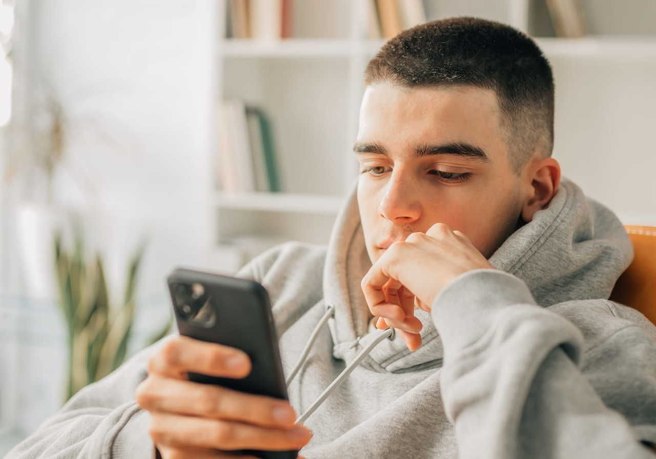 A young man in a gray hoodie sits casually, looking at his phone with a thoughtful expression.