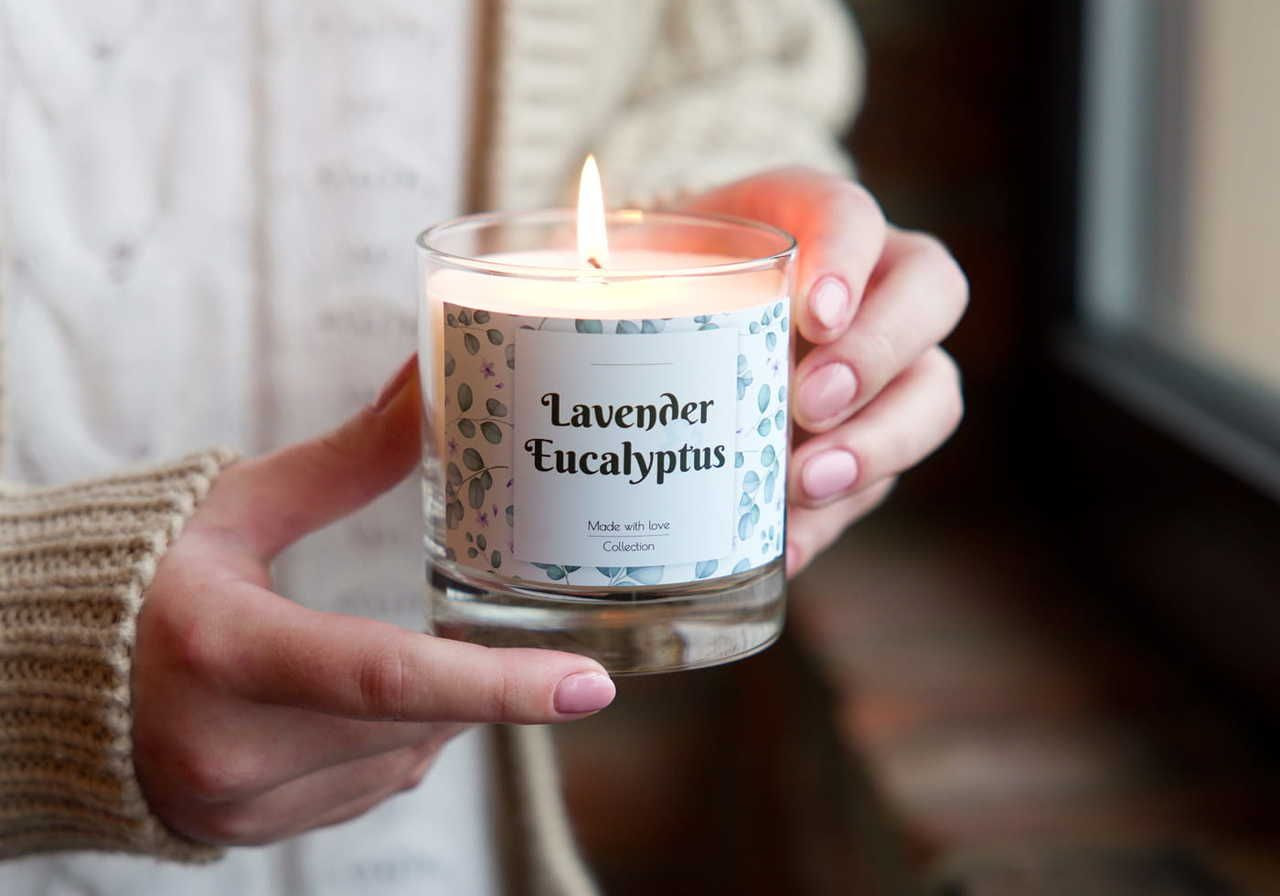 A person holding a candle labeled "Lavender Eucalyptus," with a flame lit, wearing a knitted sweater.