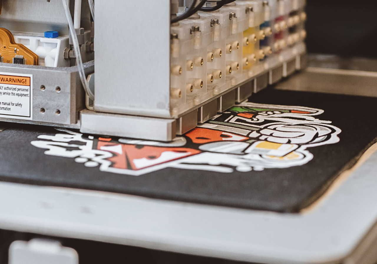 Close-up of an industrial printer applying colorful ink to fabric, featuring a vibrant cartoon design. Labels and cables visible.