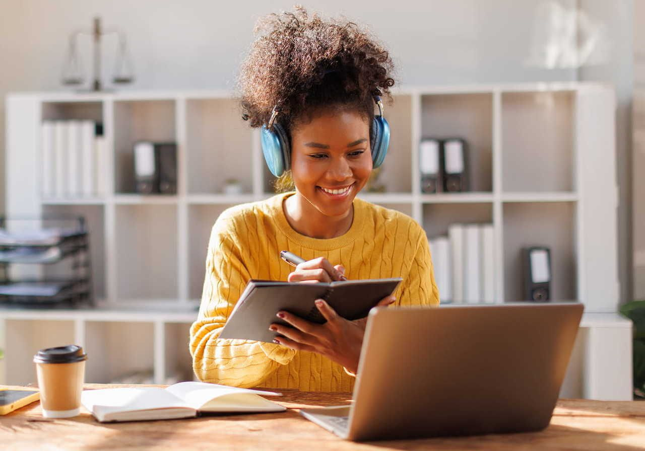 A young woman with curly hair wearing headphones, smiling while making notes and looking at a laptop.