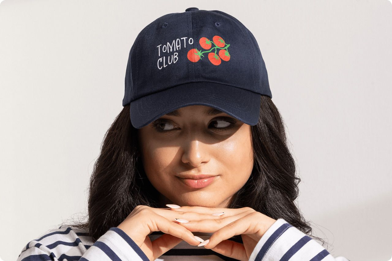 A woman wearing a hat that reads "Tomato Club," smiling and enjoying a sunny day outdoors.