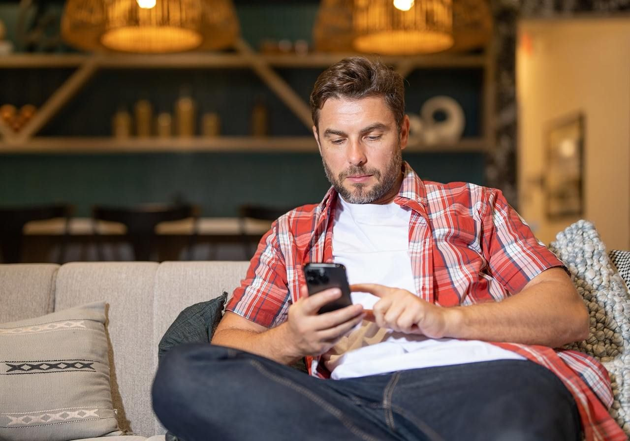 A man in a red plaid shirt sits on a couch, focused on his smartphone, searching for information on key TikTok metrics.