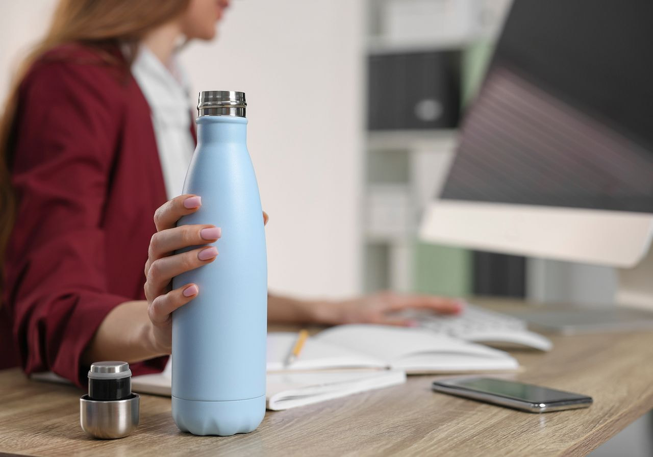 A woman stands holding a water bottle in one hand and a laptop in the other, smiling at the camera.