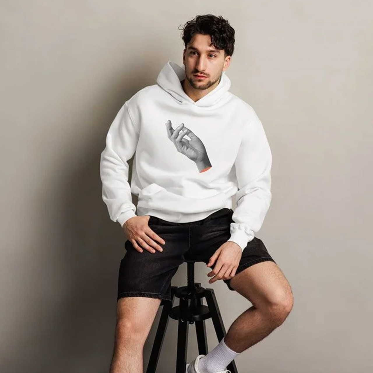 A man in a white hoodie featuring a hand graphic sits on a stool against a neutral background. He wears black shorts, white socks.