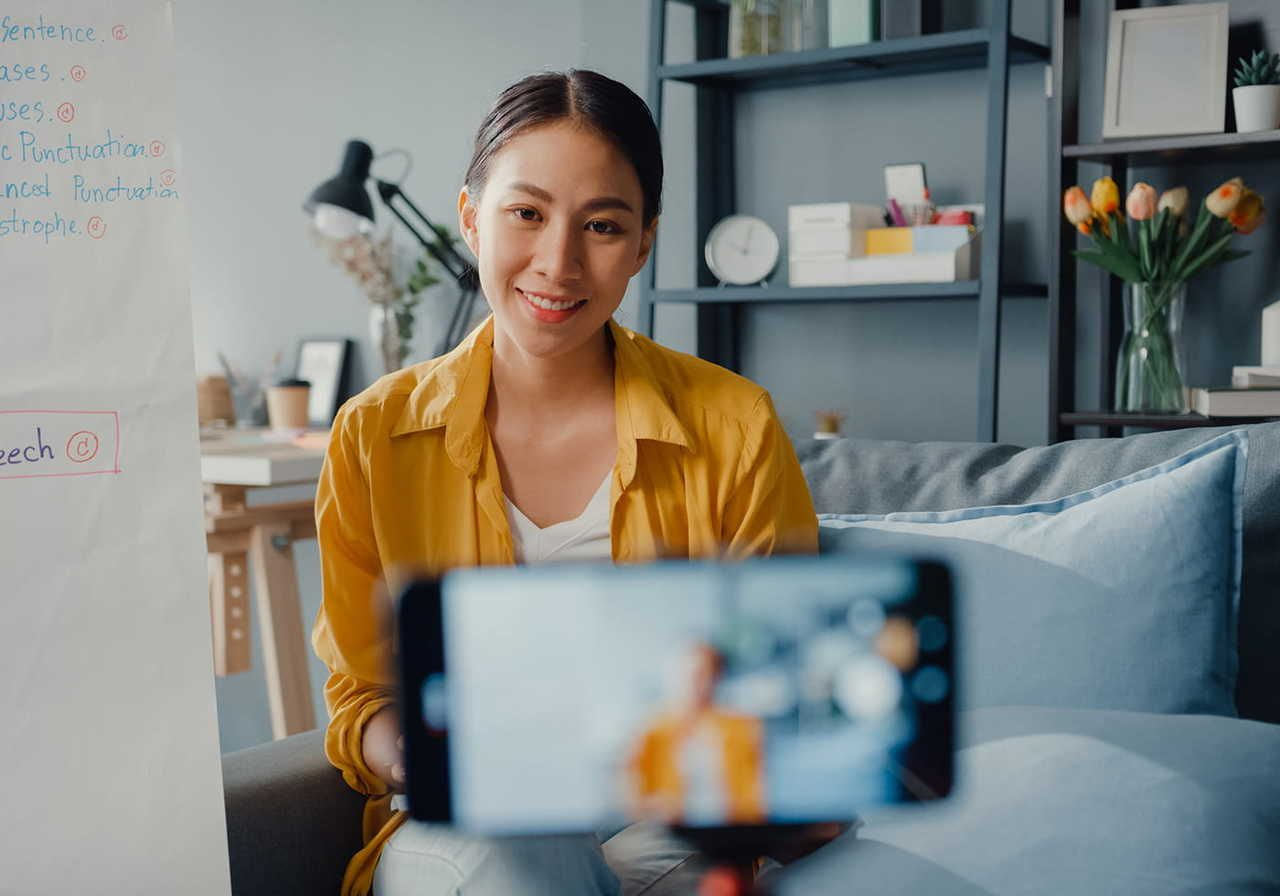 A smiling woman in a yellow blouse is recording herself with a smartphone. Home office in the background.