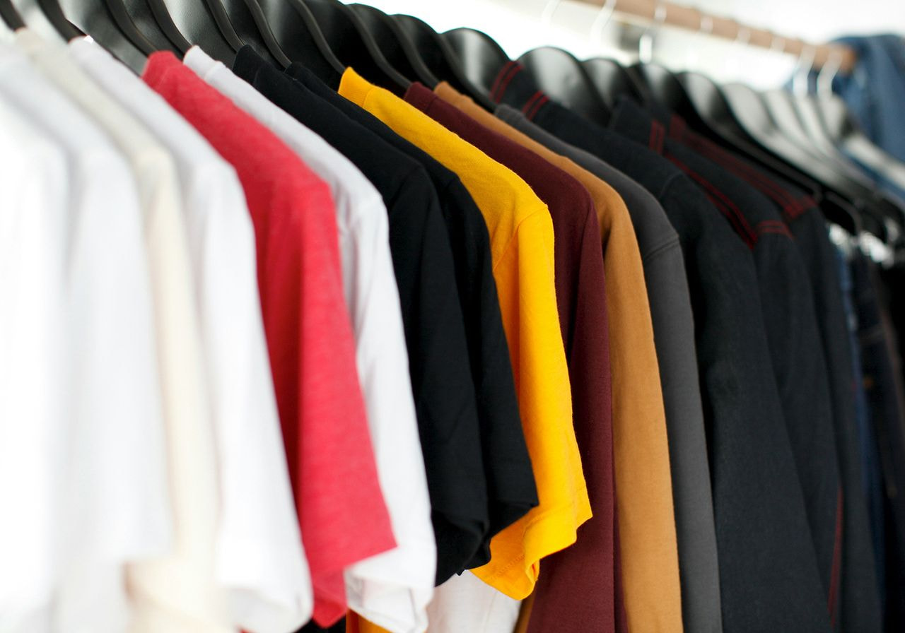 A row of t-shirts in various colors, including white, red, yellow, and black, hanging neatly on black hangers in a closet. 
