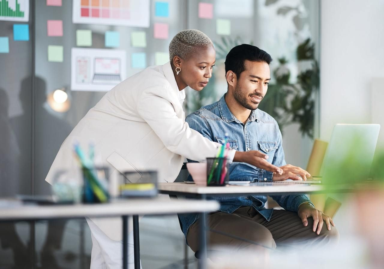 A woman in a white suit leans over a seated man in a denim shirt, pointing and focusing on his laptop.