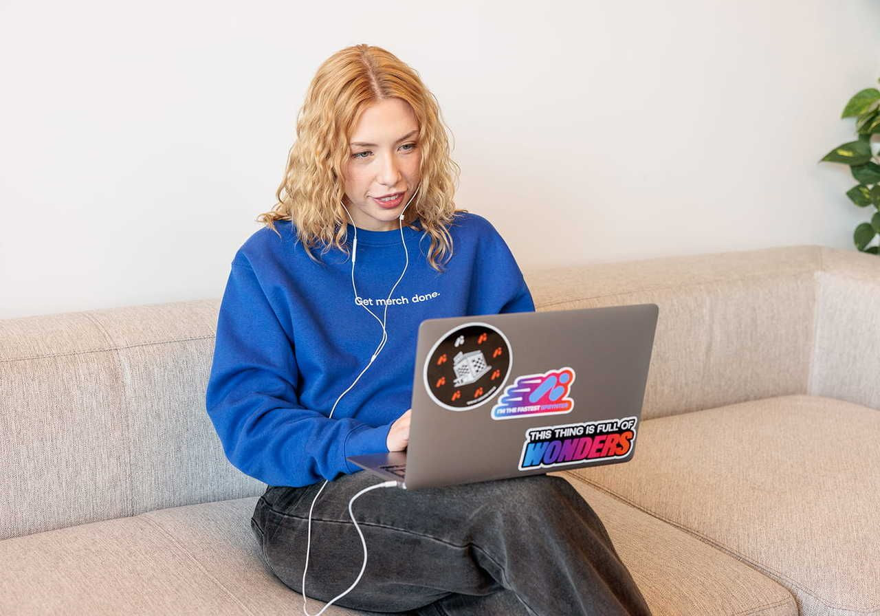 A woman with curly blonde hair is sitting on a light-colored couch, working on a laptop covered in colorful stickers.