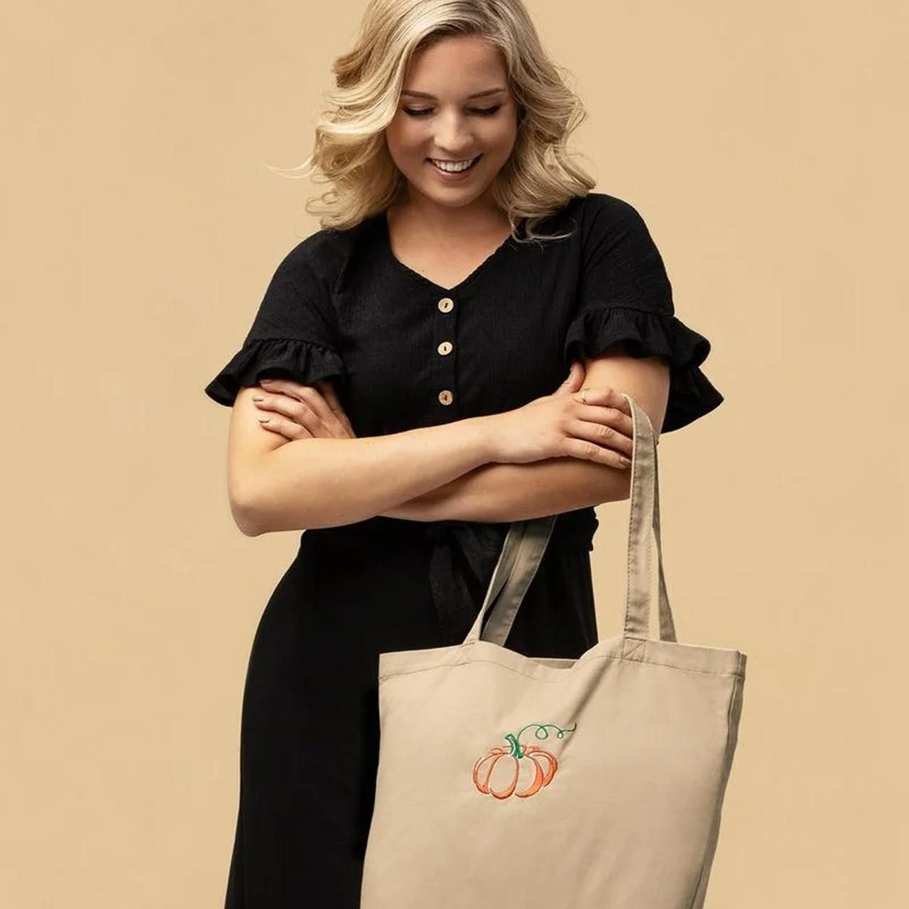 A smiling woman in a black dress holds a beige eco-friendly tote bag with an embroidered pumpkin design.
