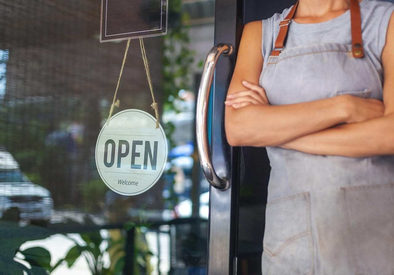 A woman stands in front of a business displaying an open sign, welcoming customers to enter.