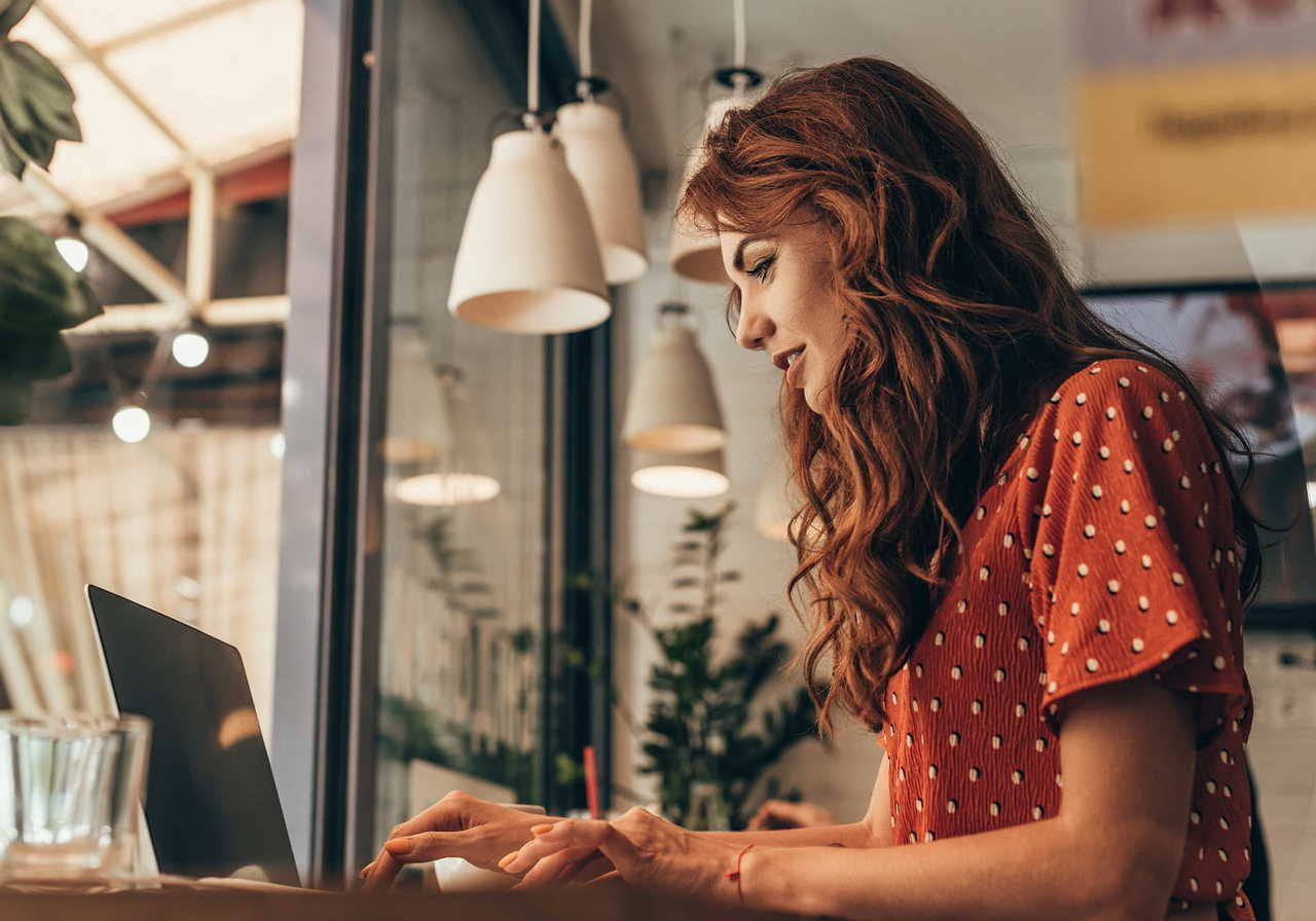 A woman with red hair, wearing a red dress with white dots, is blogging on a laptop in a cafe.