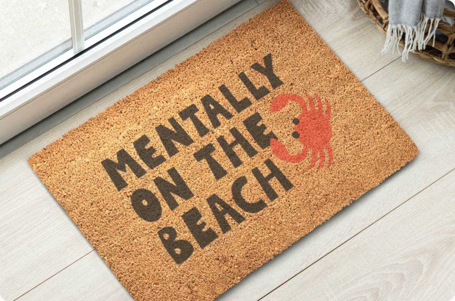 Natural coir doormat placed by a doorway, printed with a humorous beach-themed phrase and a red crab illustration.