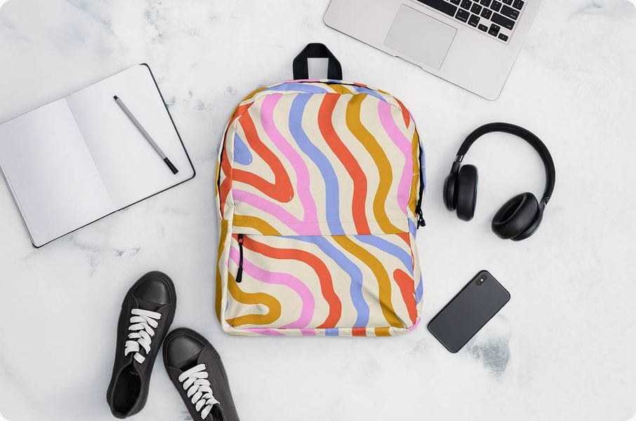 A flat lay photo showing a cream-colored backpack with a retro, abstract wavy line pattern in pink, blue, orange, and mustard yellow. The backpack is surrounded by accessories on a white marble surface: a laptop, black headphones, a smartphone, a notebook, and a pair of black sneakers.