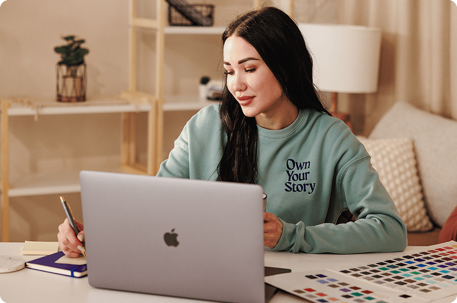 Woman working on a laptop at a home office desk while wearing a custom embroidered crewneck sweatshirt featuring the phrase “Own Your Story,” with color swatches and design materials nearby, highlighting personalized apparel creation.