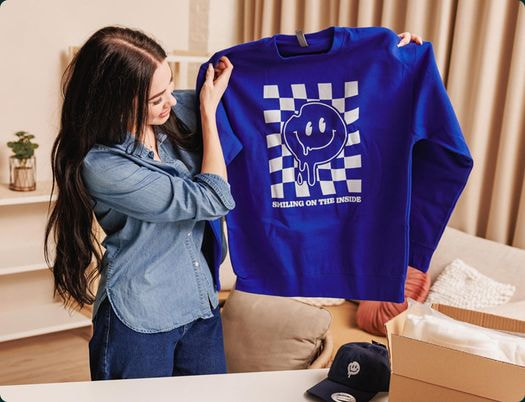 Woman holding up a bright blue sweatshirt with smiley face graphic print design.