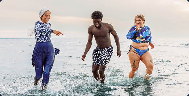 Diverse group of friends wearing custom swimwear and swim shorts while running through shallow ocean water.