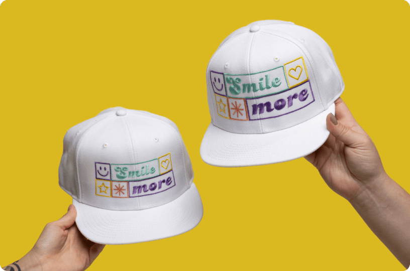 White snapback hats with colorful embroidered ‘Smile more’ design, shown being held by hands against a yellow background.