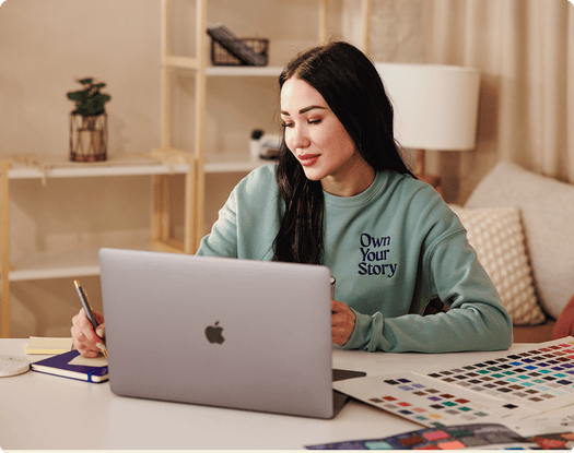 Person working on a laptop at a desk with color swatches and design samples while wearing a sweatshirt that reads Own Your Story.