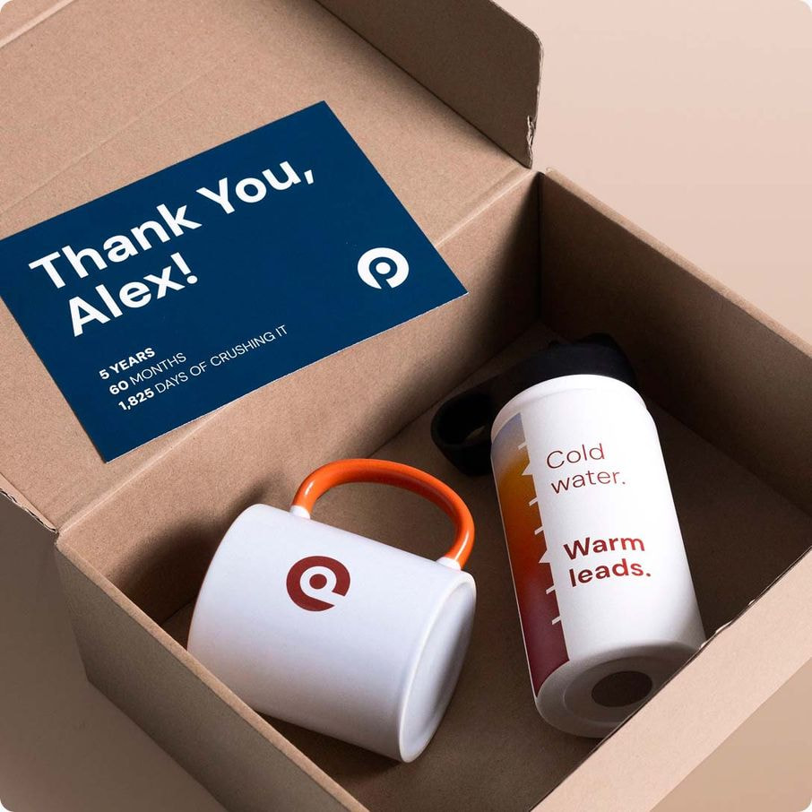 Overhead view of an open cardboard box containing a mug with an orange handle, a water bottle, and a thank-you card with a milestone message and logo.