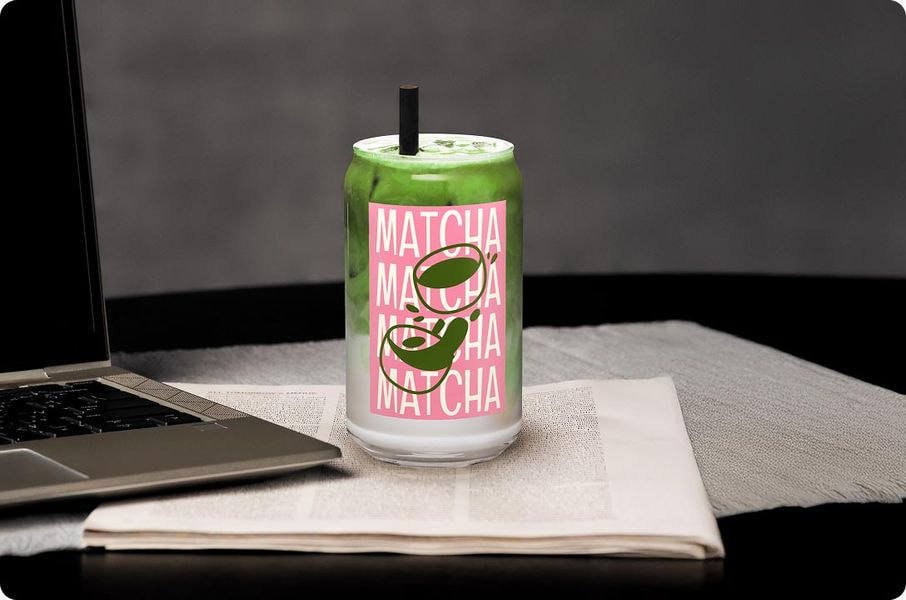 A close-up, high-quality photograph of a finished product: a clear can-shaped glass filled with a green, frothy matcha latte and a black straw. The glass has a customized label featuring the word 